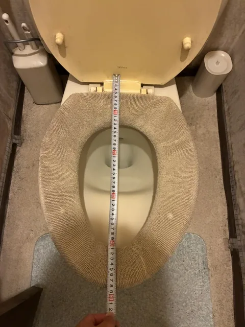 川口市のトイレ普通便座交換|施工前。便座カバーが掛かった既設の便座と、便器の大きさを測っているメジャー